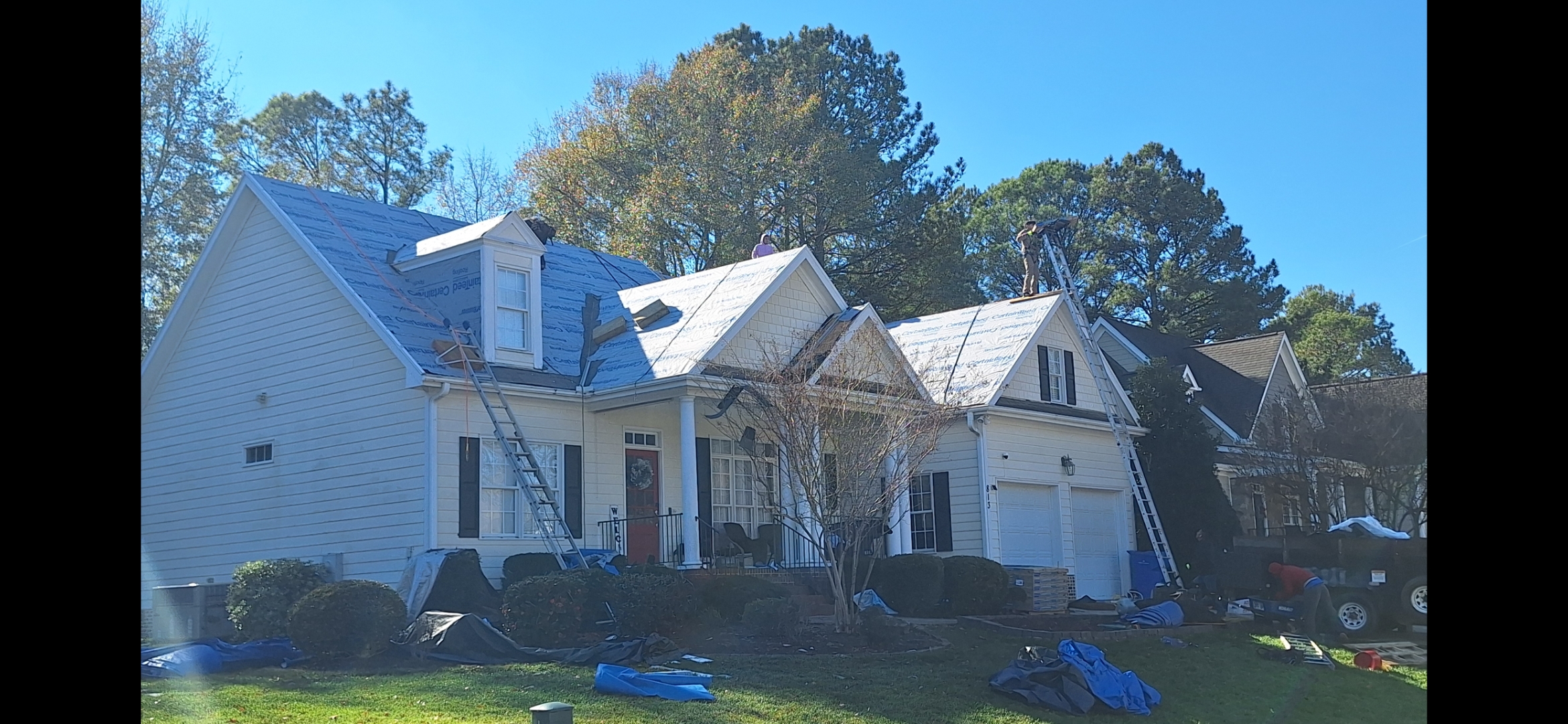 Roof replacement in progress with ladders, workers, and blue tarps around house